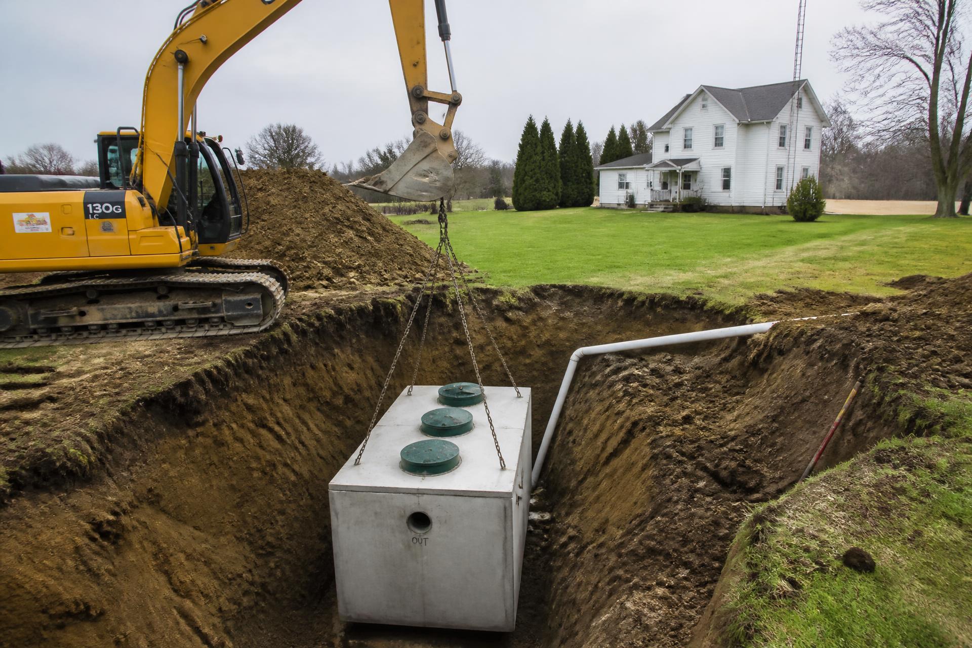 Septic system installation in Urbana Ohio: an excavator lowering a new concrete septic tank into a trench beside a rural Champaign County home